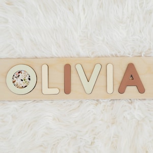 May include: Wooden puzzle with the name "OLIVIA" spelled out in individual pieces. The letters are painted in different colors, including beige, light green, and brown. A circular piece with a floral pattern is included in the puzzle.