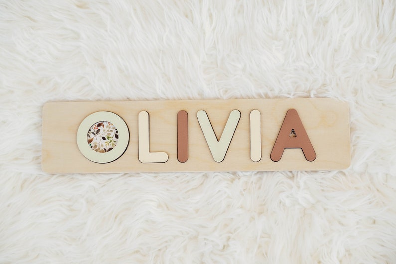 May include: Wooden puzzle with the name "OLIVIA" spelled out in individual pieces. The letters are painted in different colors, including beige, light green, and brown. A circular piece with a floral pattern is included in the puzzle.