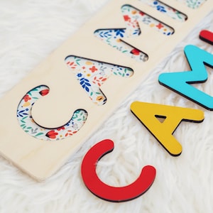 May include: Wooden name puzzle with the name "CAMILA" cut out. The letters are in various colors, with the letters "C", "A", and "M" in red, yellow, and blue. The letters are on a light wood base, and the cut-out letters have a floral pattern.