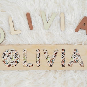 May include: Wooden puzzle with the name "OLIVIA" cut out. The letters are separate and can be placed in the cutouts. The letters are painted in various shades of brown, beige, and green. The puzzle has a floral pattern.