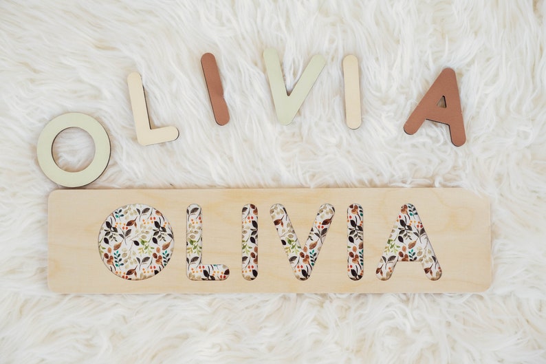 May include: Wooden puzzle with the name "OLIVIA" cut out. The letters are separate and can be placed in the cutouts. The letters are painted in various shades of brown, beige, and green. The puzzle has a floral pattern.