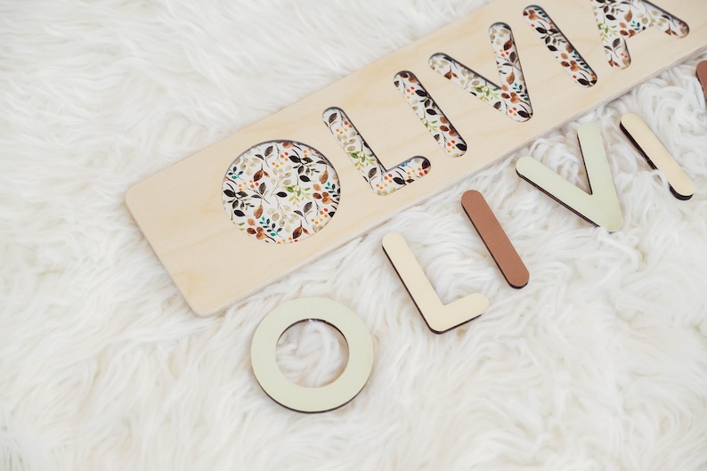 May include: Wooden puzzle with the name "Olivia" cut out. The letters are separate and can be placed in the cutouts. The background of the puzzle is a light wood with a floral pattern.