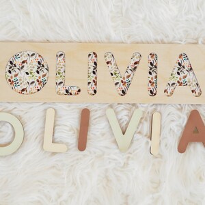 May include: Wooden puzzle with the name "OLIVIA" cut out. The letters are painted in various shades of brown and beige and are arranged on a white fuzzy surface. The puzzle board has a floral pattern.