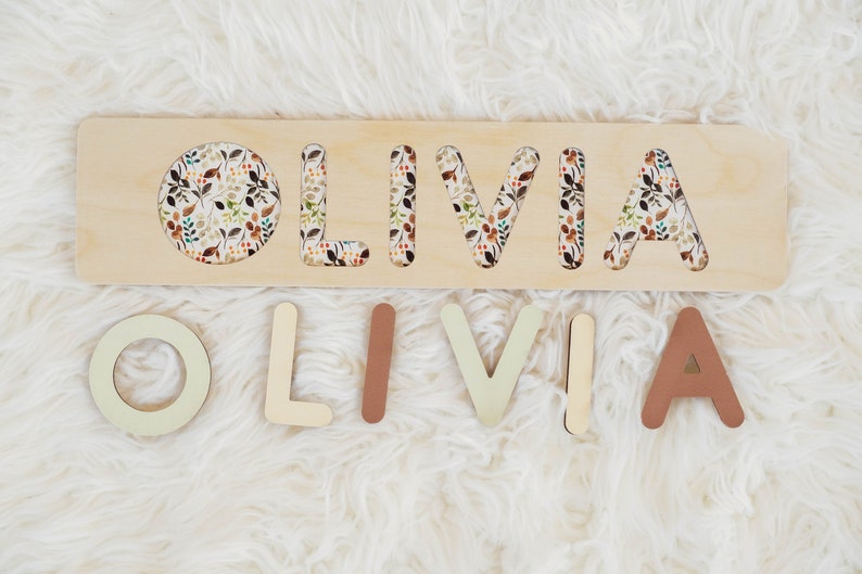 May include: Wooden puzzle with the name "OLIVIA" cut out. The letters are painted in various shades of brown and beige and are arranged on a white fuzzy surface. The puzzle board has a floral pattern.