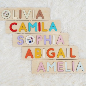 May include: Five wooden blocks with the names Olivia, Camila, Sophia, Abigail, and Amelia spelled out in colorful letters. Each letter is a different color and is raised from the surface of the block.