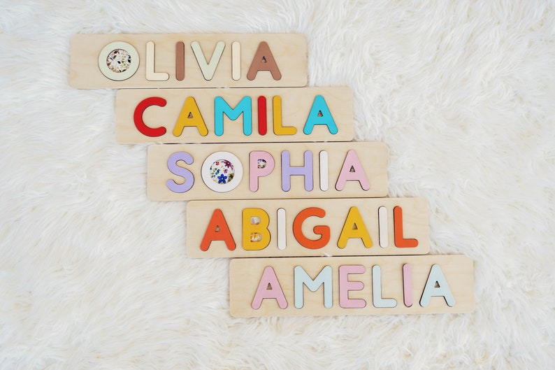 May include: Five wooden blocks with the names Olivia, Camila, Sophia, Abigail, and Amelia spelled out in colorful letters. Each letter is a different color and is raised from the surface of the block.