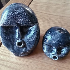 May include: Two ceramic face sculptures with a dark blue speckled glaze. The larger sculpture is oval-shaped, and the smaller one is round. Both have stylized facial features, including closed eyes and open mouths.