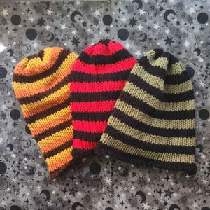 May include: Three knitted beanies in a row. The first beanie has horizontal stripes of orange, yellow, and brown. The second beanie has red and black stripes. The third beanie has black and olive green stripes. The beanies are on a gray background with a celestial pattern.