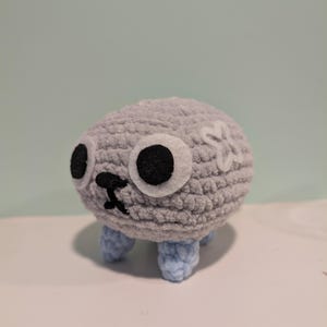 Puede incluir: Un juguete de peluche gris tejido a crochet con ojos negros, una nariz negra y una flor blanca en la cabeza. El juguete tiene cuatro patas azules tejidas a crochet.