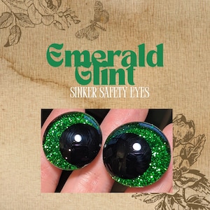 May include: Two green glitter safety eyes with black pupils. The eyes are 1 inch in diameter and have a shiny finish. The text "Emerald Tint Sinker Safety Eyes" is visible above the eyes.