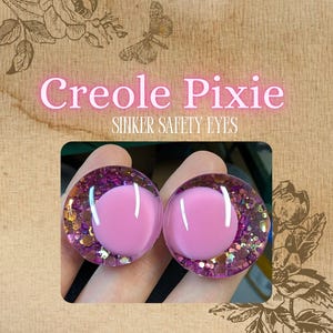May include: Two Creole Pixie sinker safety eyes. Each eye is a clear, round dome with a light pink center and a surrounding border of purple glitter and gold flecks. The background is a textured, sepia-toned paper.