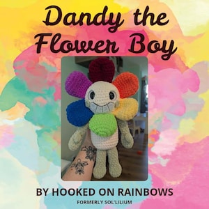 Op de afbeelding: Gehaakte bloemenpop met een lachend gezicht en kleurrijke bloemblaadjes. De bloemblaadjes zijn in de tinten rood, paars, oranje, geel, blauw en groen. De tekst "Dandy the Flower Boy" staat bovenaan en "By Hooked on Rainbows" onderaan.