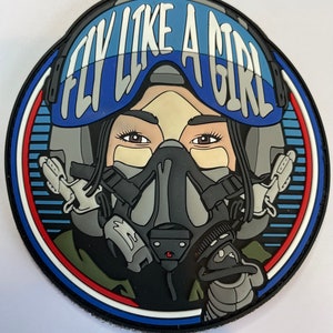 Könnte beinhalten: Ein runder schwarzer und blauer Patch mit weißem Rand und roten und weißen Streifen. Der Patch zeigt eine Frau, die einen blauen Helm mit Visier und eine Sauerstoffmaske trägt. Der Text "FLY LIKE A GIRL" ist auf dem Helm aufgedruckt.