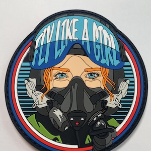 May include: A patch with a graphic of a woman in a pilot's helmet and oxygen mask. The text "FLY LIKE A GIRL" is displayed across the top. The patch is framed by a blue, red, and white border.
