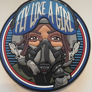 May include: Circular patch featuring a pilot with brown skin, dreadlocks, and a flight helmet. The helmet has the text "FLY LIKE A GIRL" in white. The patch has a blue, red, and white border, with a black background.