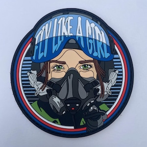 Puede incluir: Un parche moral circular de PVC con una ilustración estilizada de un piloto con casco de vuelo y máscara de oxígeno. El texto FLY LIKE A GIRL está impreso en azul claro sobre la visera azul del casco. El diseño incluye ojos verdes.