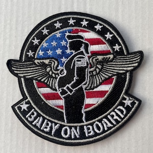 Könnte beinhalten: Ein gestickter Patch in Schwarzweiß mit einer Silhouette einer schwangeren Person in Militäruniform mit Flügeln. Die Person steht vor einer amerikanischen Flagge mit dem Text "BABY ON BOARD" in einem weißen Kreis um die Flagge.