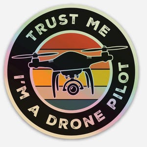 Peut inclure: Un autocollant rond noir et blanc avec un effet de dégradé arc-en-ciel. L'autocollant représente un drone avec une caméra, un fond de coucher de soleil et le texte "Trust Me I'm A Drone Pilot".