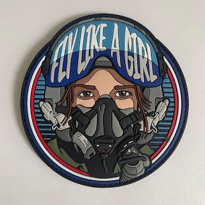 May include: A circular PVC morale patch featuring a pilot wearing a flight helmet and oxygen mask. The text FLY LIKE A GIRL is printed in white across the blue helmet visor. The design includes shades of blue, black, gray, and red accents.