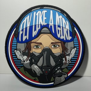 May include: A round, black, white, red, and blue embroidered patch with a woman wearing a pilot's helmet and oxygen mask. The text "FLY LIKE A GIRL" is written in white letters on a blue background.