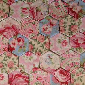 May include: A collection of fabric hexagons arranged in a mosaic pattern, featuring various floral prints in shades of pink, red, cream, and light blue. Each individual hexagon displays distinct botanical motifs, including roses and small sprigs.