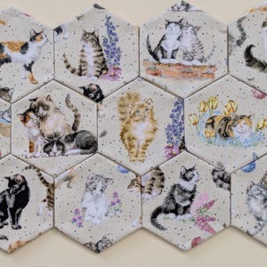 May include: A collection of ten hexagonal ceramic tiles featuring various cat illustrations. Each tile displays different feline breeds in diverse poses, including sitting, playing, and resting, accented with floral elements and a speckled cream background.