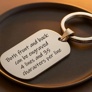 May include: A silver dog tag keychain with a polished finish, resting on a dark brown plate. The tag has the text: "Both front and back can be engraved 4 lines and 35 characters per line."