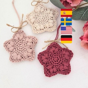 Könnte beinhalten: Drei handgefertigte Häkelstern-Ornamente in verschiedenen Rosa- und Beigetönen. Jeder Stern hat ein strukturiertes Muster und ist mit einer Schnurschlaufe zum Aufhängen versehen. Die Sterne werden auf einer weißen Oberfläche präsentiert.