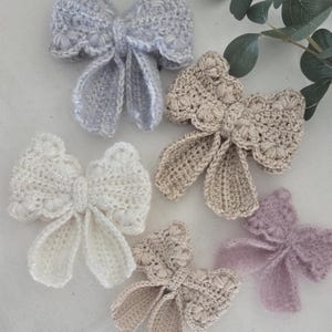 May include: A collection of handmade crochet bows in various pastel colors, including light blue, cream, beige, and lavender. Each bow features a detailed, textured design with delicate scalloped edges, showcasing the intricate craftsmanship. These bows are ideal for hair accessories or embellishments.
