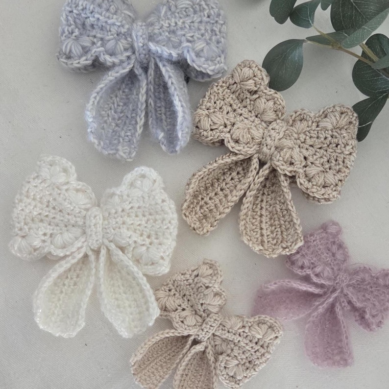 May include: A collection of handmade crochet bows in various pastel colors, including light blue, cream, beige, and lavender. Each bow features a detailed, textured design with delicate scalloped edges, showcasing the intricate craftsmanship. These bows are ideal for hair accessories or embellishments.