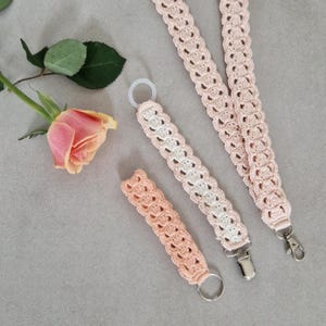 Op de afbeelding: Een verzameling handgemaakte haakartikelen in zacht perzik en wit. De set bevat een fopspeenclip, een sleutelhanger en twee lanyards met metalen sluitingen. Er is ook een roze roos met groene bladeren aanwezig.