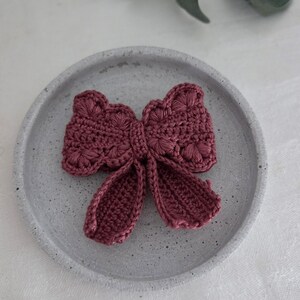 May include: A handmade, crocheted bow in a deep rose color, resting on a gray, textured plate. The bow features intricate stitch detailing and long, flowing tails. The craft item is a decorative accessory.