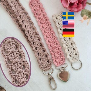 May include: Three handmade crochet lanyards in shades of taupe, pink, and cream. Each lanyard features an openwork design with a metal clasp or ring. The lanyards are displayed on a white surface with a floral background.