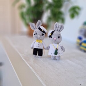 May include: Two crocheted gray bunny figurines wearing sailor hats. The bunny on the left is wearing a yellow bow tie and black pants. The bunny on the right is wearing a white dress with a green pendant.