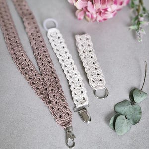 Op de afbeelding: Gehaakte lanyards en accessoires in tinten taupe en wit. De lanyards hebben een decoratief opengewerkt patroon en worden getoond met een metalen sluiting, een fopspeenclip en een sleutelring. De items worden gepresenteerd op een grijze ondergrond.