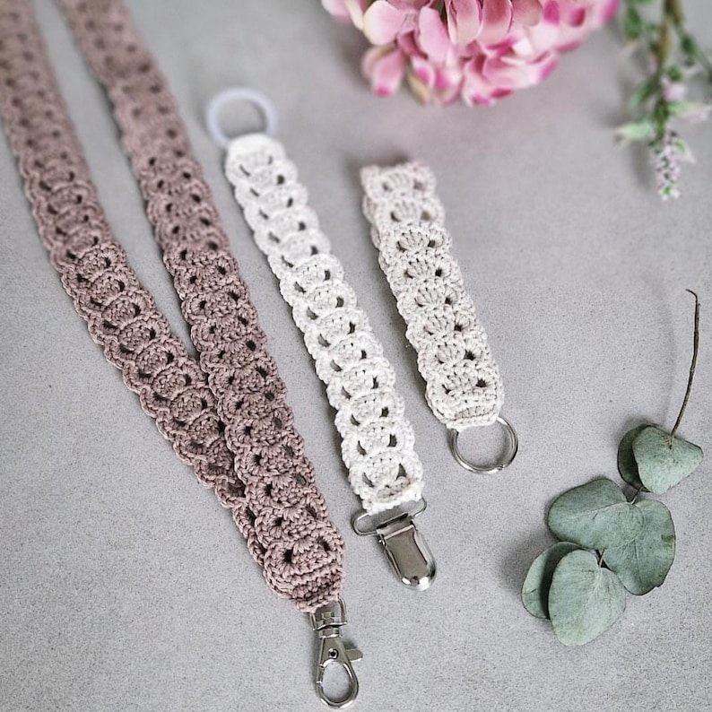 Op de afbeelding: Gehaakte lanyards en accessoires in tinten taupe en wit. De lanyards hebben een decoratief opengewerkt patroon en worden getoond met een metalen sluiting, een fopspeenclip en een sleutelring. De items worden gepresenteerd op een grijze ondergrond.