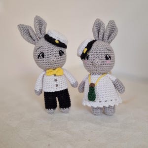 May include: Two gray crocheted bunny plushes wearing sailor hats. One bunny is wearing a white shirt with a yellow bow tie and black pants. The other bunny is wearing a white dress with a green bottle charm.
