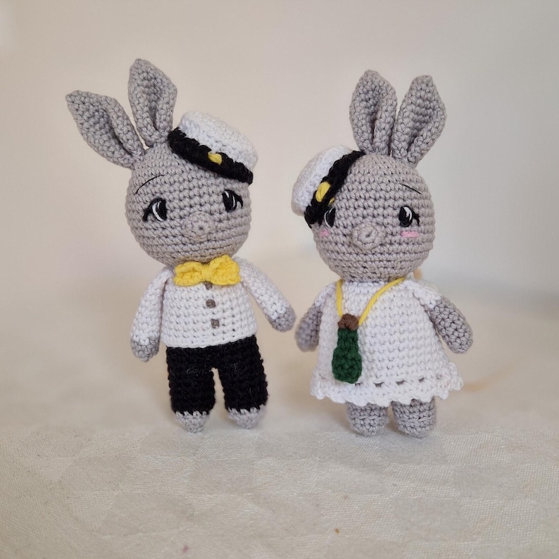 May include: Two gray crocheted bunny plushes wearing sailor hats. One bunny is wearing a white shirt with a yellow bow tie and black pants. The other bunny is wearing a white dress with a green bottle charm.