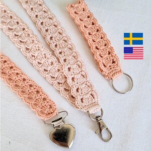 Op de afbeelding: Vier handgemaakte gehaakte lanyards in perzik- en cr&egrave;metinten. De lanyards hebben een herhalend opengewerkt patroon en zijn afgewerkt met metalen hardware, waaronder een hartvormige clip, een draaibare sluiting en een splitring. De lanyards worden op een wit oppervlak gepresenteerd.