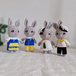 May include: Four crocheted gray bunny plushies wearing different outfits. One bunny is wearing a blue and yellow dress, another is wearing a blue and yellow jumpsuit, another is wearing a white dress, and the last one is wearing a white shirt, black pants, and a black and white sailor hat.