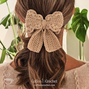 May include: A tan crochet bow hair accessory. The bow is attached to a ponytail, with the back of a person's head and shoulders visible. The bow is made of a textured yarn and has a detailed floral pattern. The text "Coffee n Crochet" is in the lower right corner.