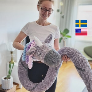 May include: A woman holds a large crocheted unicorn pillow. The pillow is gray and purple with a white horn. The unicorn is curled into a circle.