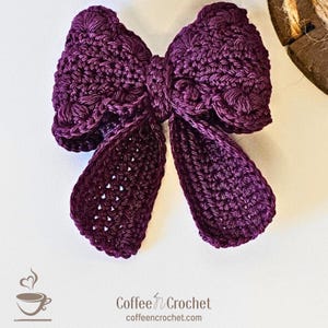 May include: A handmade, dark purple crochet bow. The bow features a detailed, textured stitch pattern and long, flowing tails. The image also includes the text "Coffee n Crochet" and a coffee cup logo.