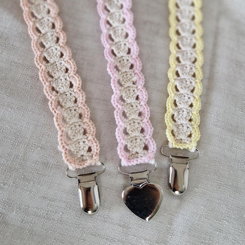 Op de afbeelding: Drie gehaakte fopspeenclips in pastelkleuren: roze, perzik en geel. Elke clip heeft een decoratief haakpatroon en een metalen clip. E&eacute;n clip heeft een hartvormige clip.