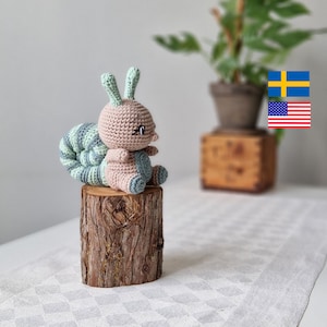 Puede incluir: Un juguete de caracol de crochet con un cuerpo marrón, una concha rayada de verde y blanco y ojos negros. El caracol está sentado en un tocón de árbol.