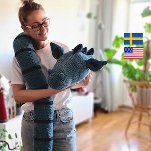 Puede incluir: Una mujer sostiene un gran juguete de dragón de crochet azul y negro. El dragón tiene un cuerpo largo y delgado y una cabeza grande con una expresión amigable. La mujer sonríe y mira a la cámara.