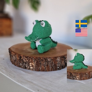 May include: A green crocheted alligator toy sitting on a wooden slice. The alligator has white eyes and a white belly.