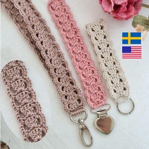 Op de afbeelding: Gehaakte polsbandjes in roze en beige tinten, met een decoratief openwerkpatroon. Elke band heeft een metalen sluiting of ring voor bevestiging. De afbeelding bevat een close-up van een enkele band en een bloem op de achtergrond.