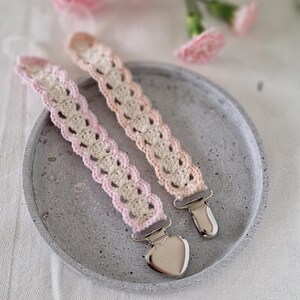 Op de afbeelding: Twee gehaakte fopspeenclips in roze en cr&egrave;me. Elke clip heeft een geschulpte rand en een metalen clip. E&eacute;n clip heeft een hartvormige clip, de andere een rechthoekige. De clips liggen op een grijze plaat.