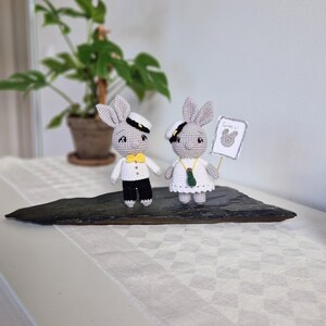 May include: Two crocheted bunny figurines dressed in sailor outfits. The bunny on the right is holding a sign that says "Grattis!" in white lettering on a light blue background.